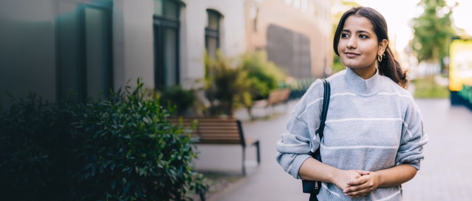 A woman in a grey striped sweater standing on a tree-lined city street, looking to one side